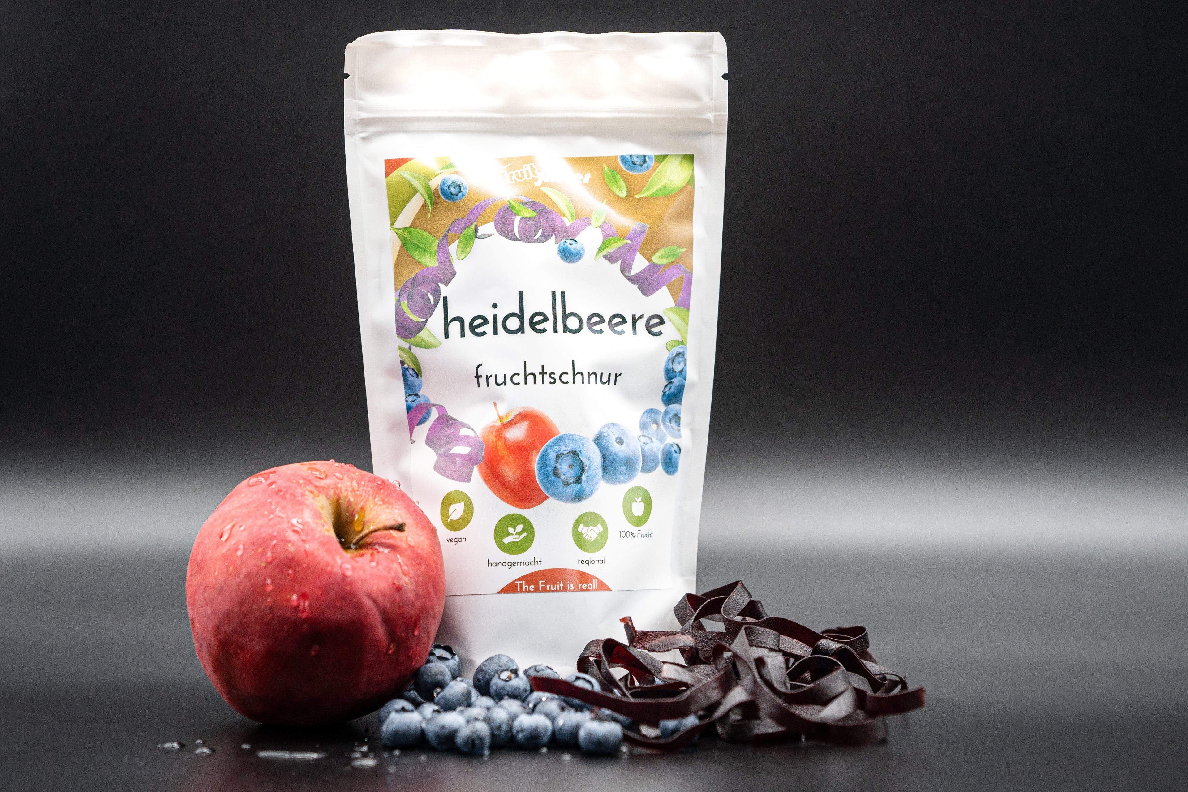 Tüte Fruchtschnur Heidelbeere und Heidelbeeren sowie ein Apfel und Fruchtschnüre vor der Tüte