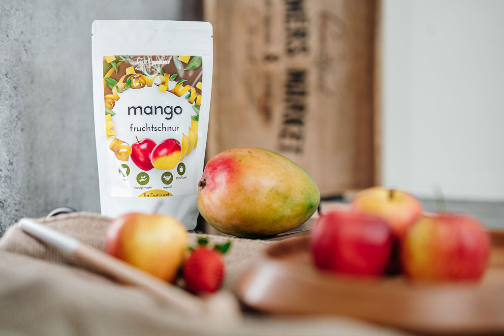 Fruchtschnurtüten Geschmacksrichtung Mango und im Vordergrund verschiedene Früchte alles auf einem Schneidbrett