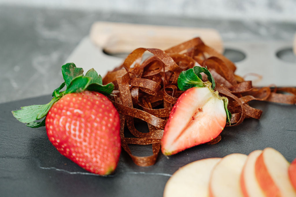 Auf einer Schieferplatte liegen Erdbeeren, Fruchtschlangen Sorte Erdbeere sowie Apfelschnitze und im Hintergrund ein Wiegemesser