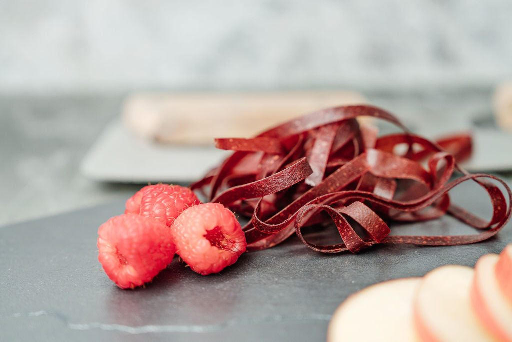 Auf einer Schieferplatte liegen Himbeeren, Fruchtschlangen Sorte Himbeere sowie Apfelschnitze und im Hintergrund ein Wiegemesser
