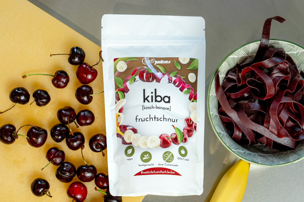 36g Tüte KiBa fruitjunkies auf einem Schneidbrett als Unterlage, rechts daneben eine Schale mit Fruchtschnüren Sauerkirsche, auch eine Banane sieht man.
