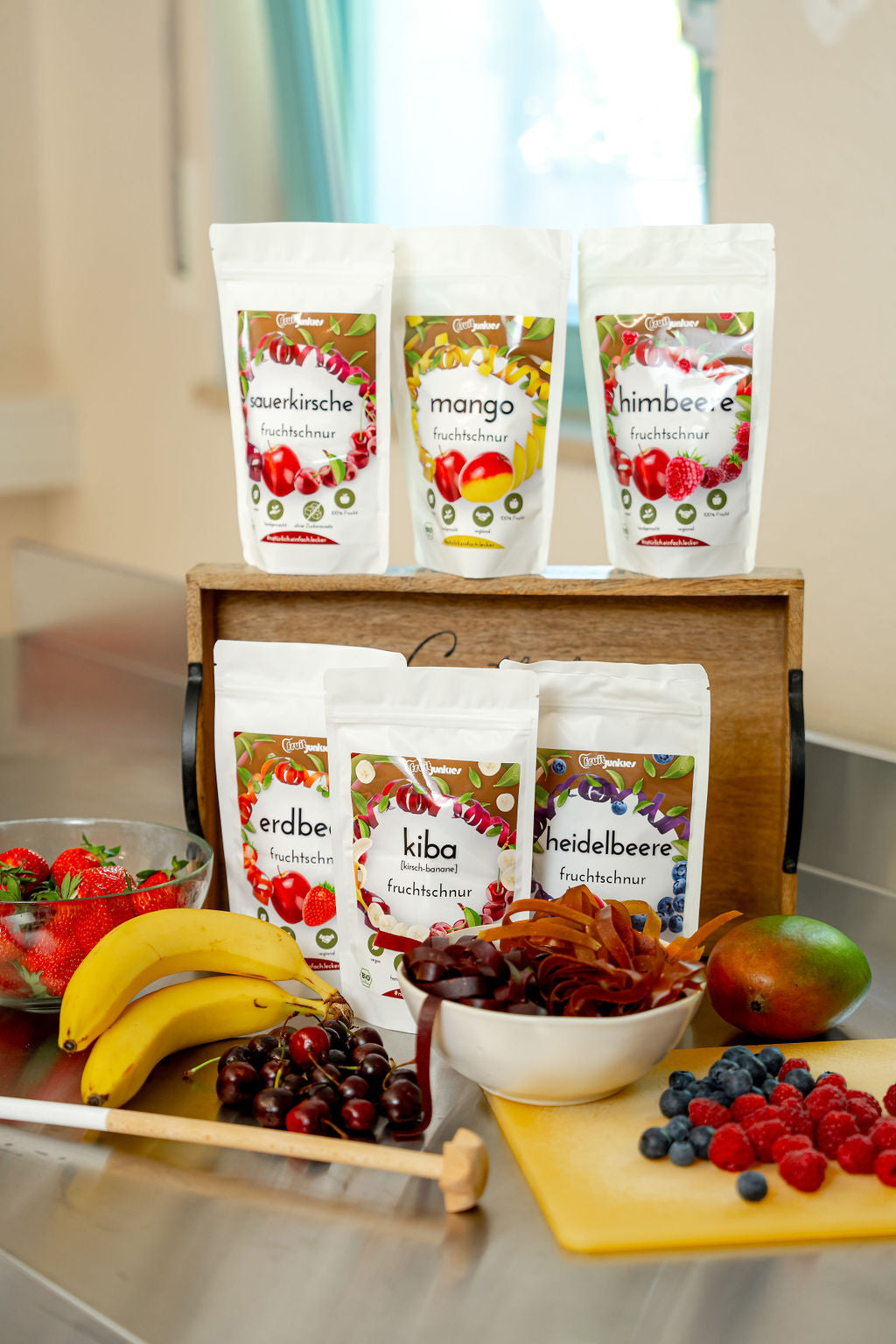 36g Tüten Fruchtschnüre von fruitjunkies in den Sorten Sauerkirsche, Mango, Himbeere, Erdbeere, KiBa, Heidelbeere. Umliegend sieht man Früchte