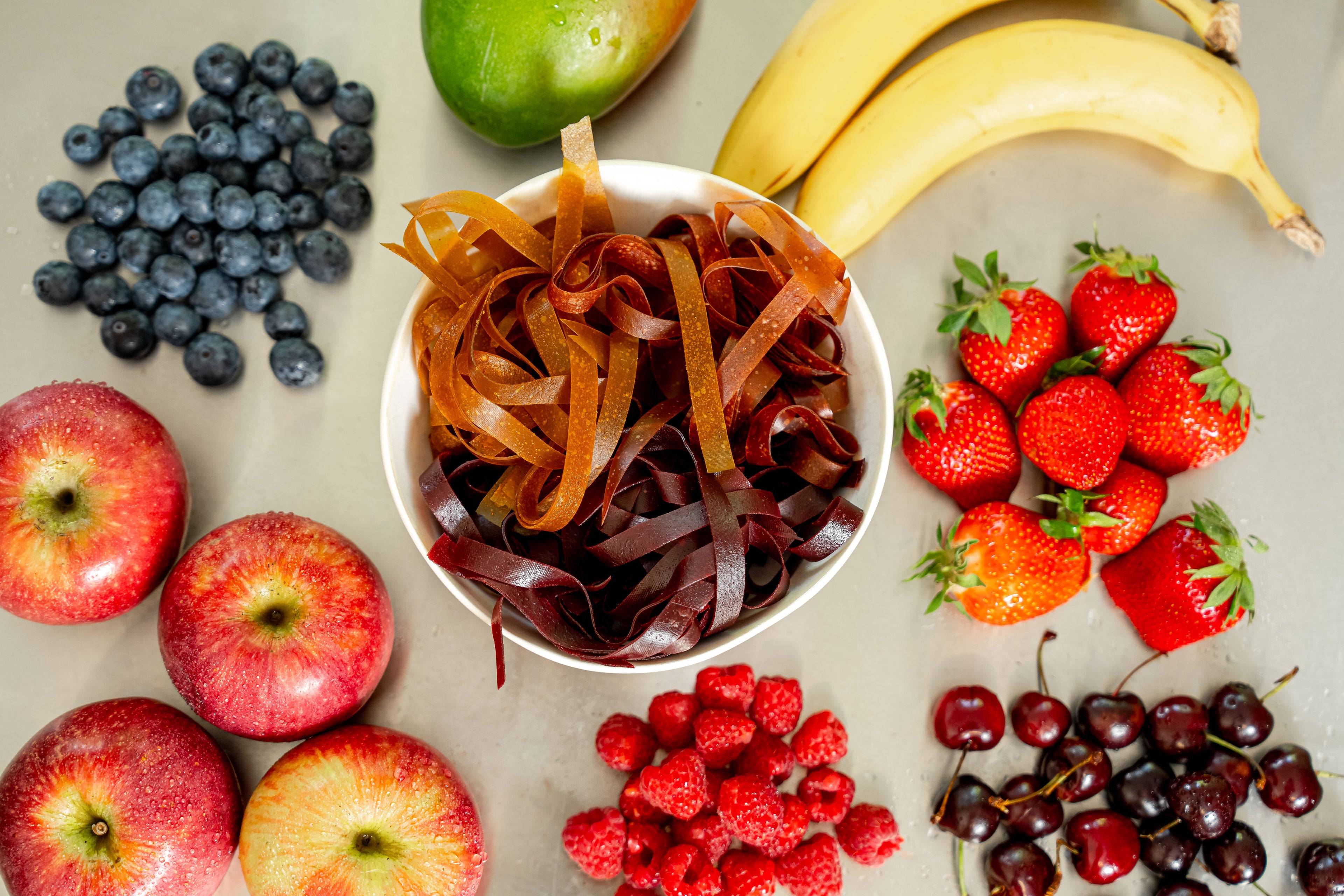 In der Mitte eine Schale mit verschiedenen Sorten Fruchtschnüren. Rundherum liegen die enthaltenen Früchte wie Bananen, Erdbeeren, Sauerkirschen, Himbeeren, Äpfel, Heidelbeere, Mango.
