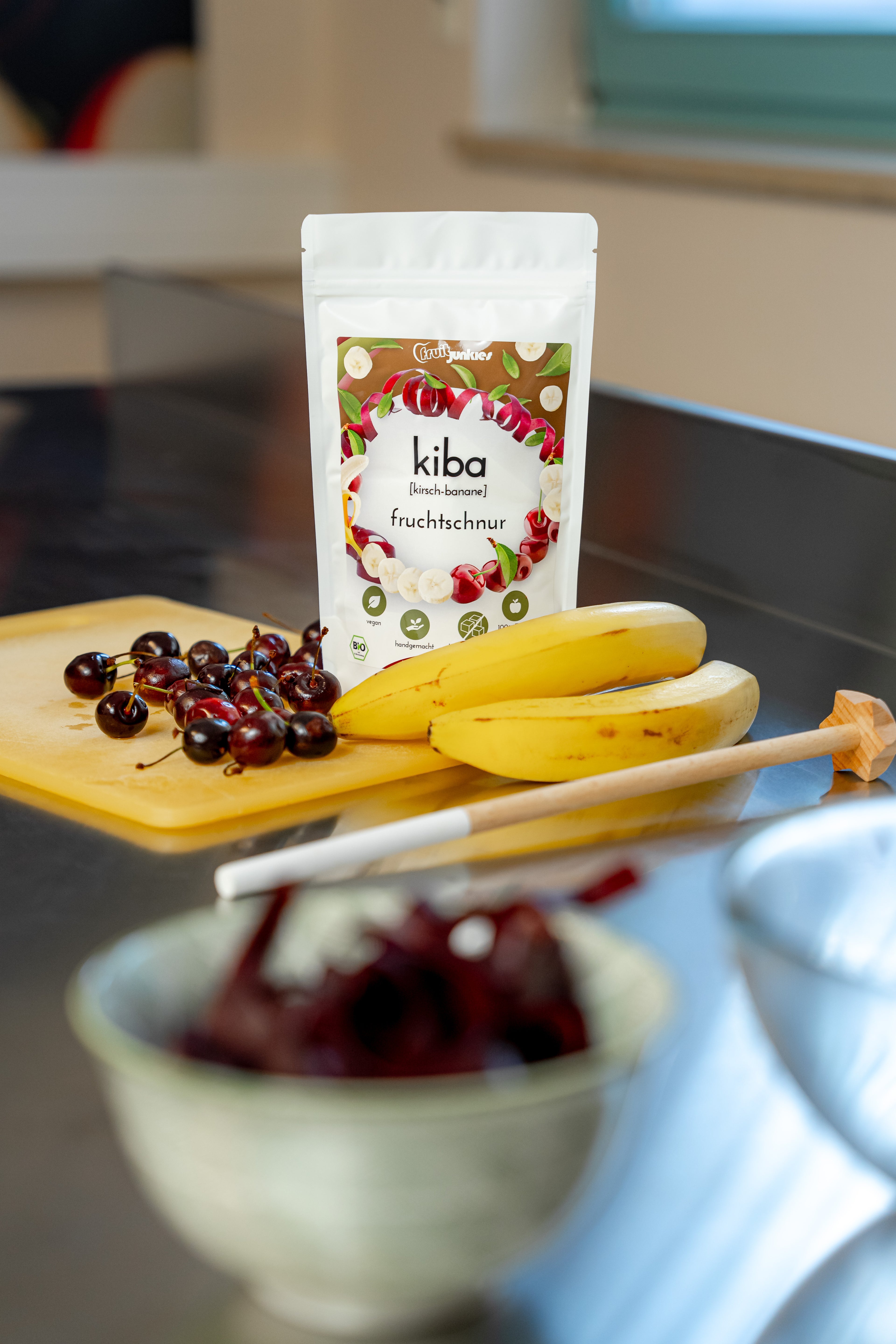 36g Fruchtschnurtüte Sorte KiBa auf einem Schneidbrett mit Bananen und Sauerkirschen und Küchenutensilien. Im Vordergrund unscharf sieht man eine Schale mit Fruchtschlangen